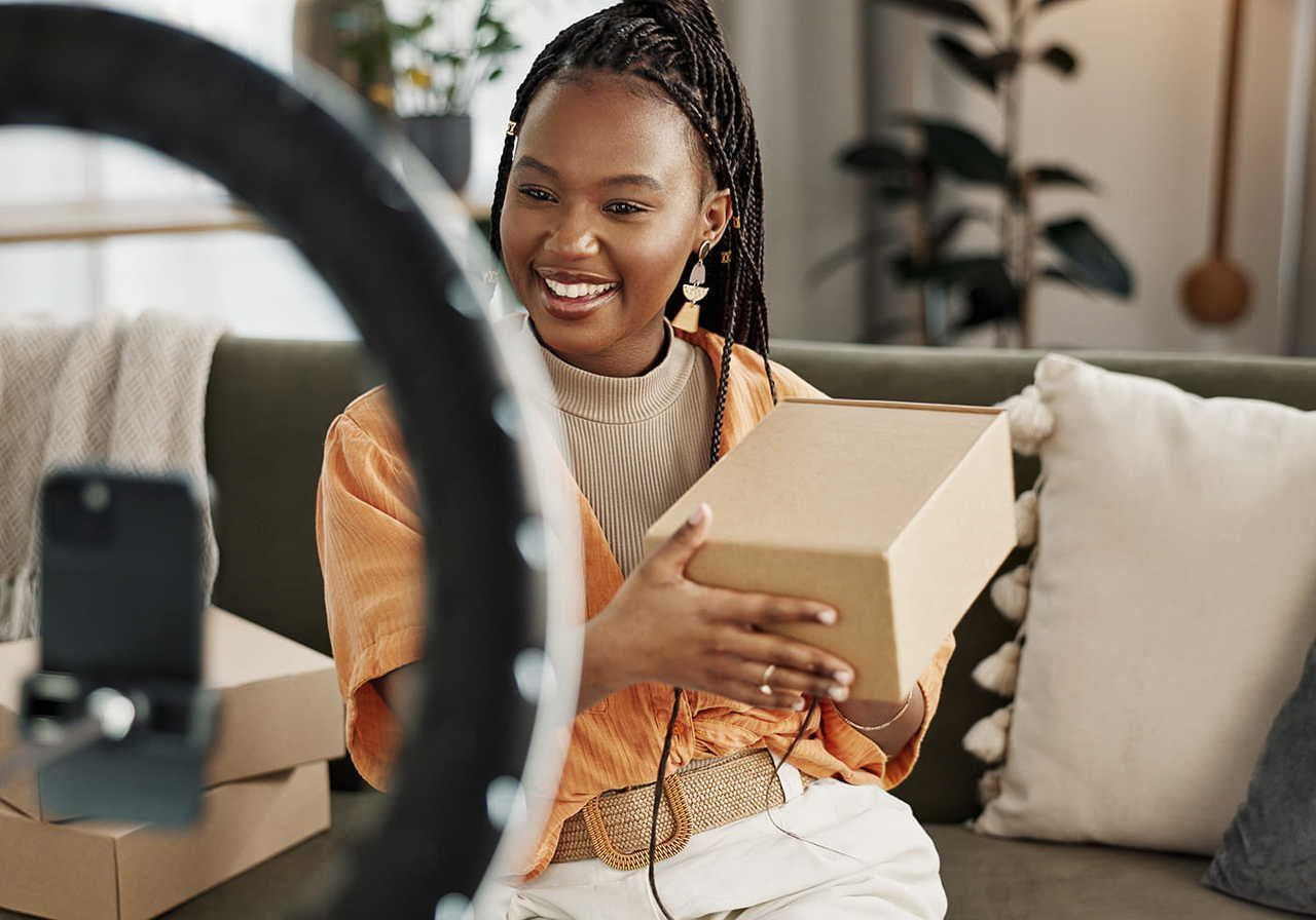 A woman sitting on a couch, smiling, holding a brown box, recording a video of herself on a smartphone with a ring light.