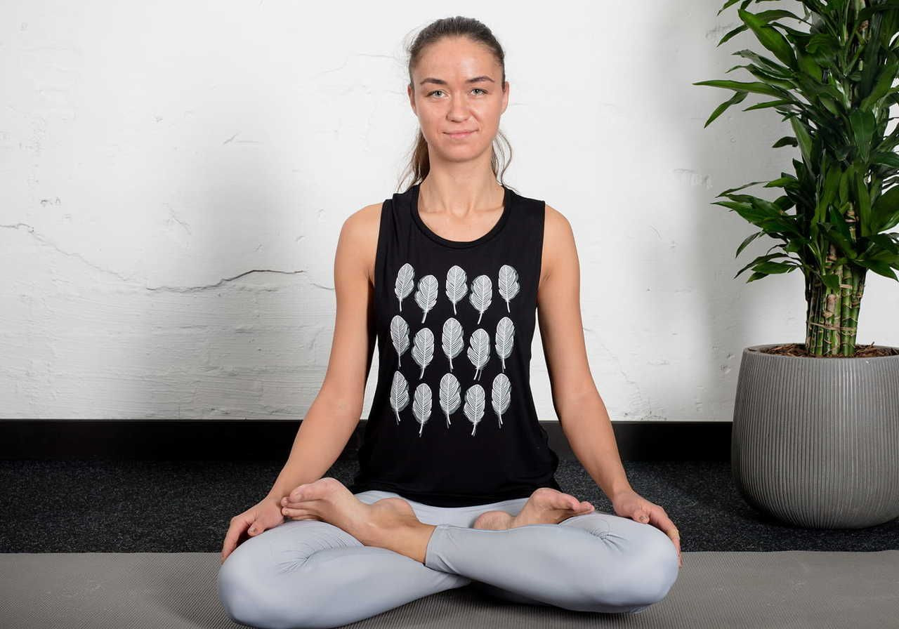 A person sitting in a yoga pose with legs crossed, wearing a black tank top with a design on it, and light gray leggings.
