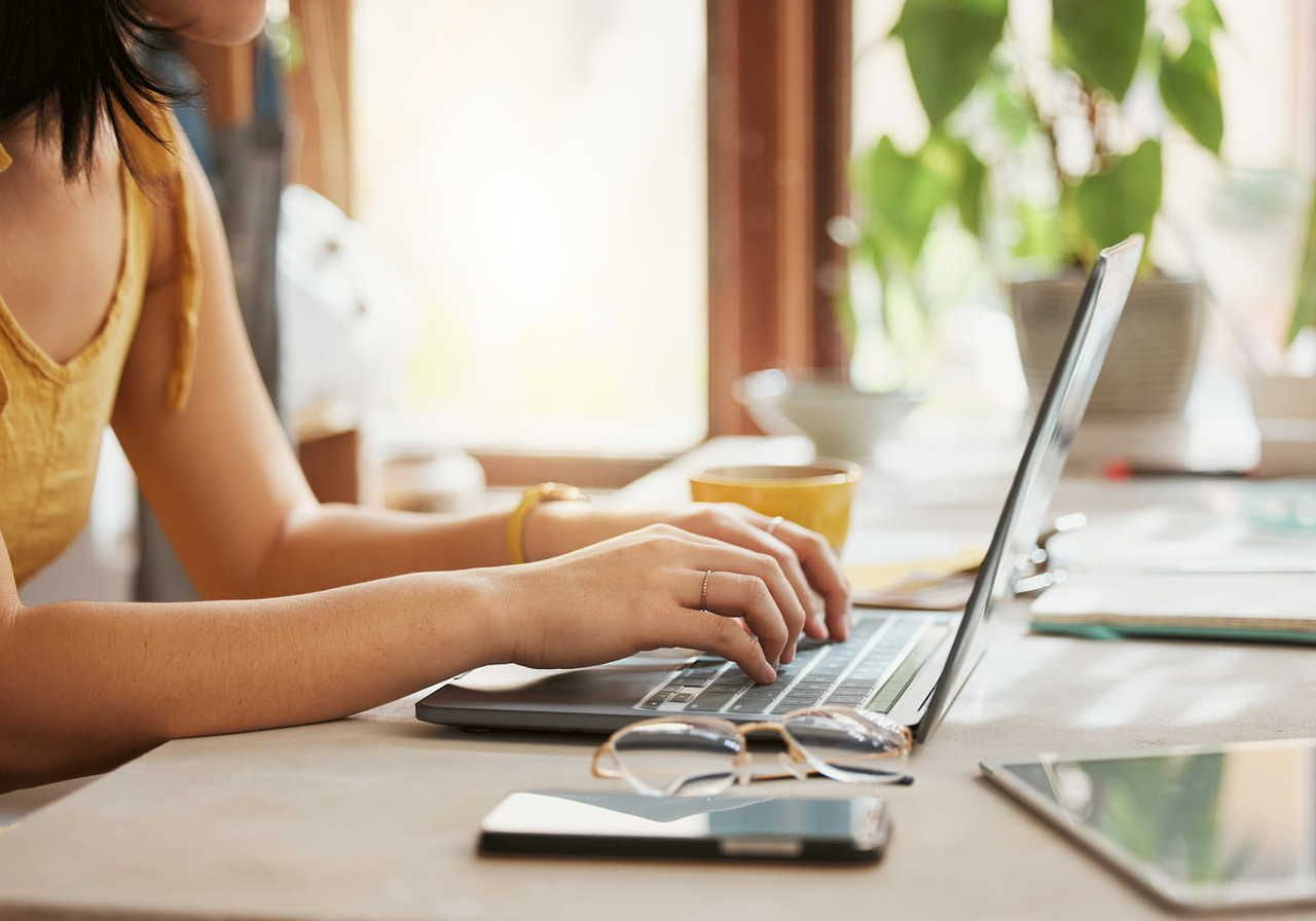 A woman is typing on a laptop keyboard to learn how to make money with print-on-demand. Blurred green plant in the background.