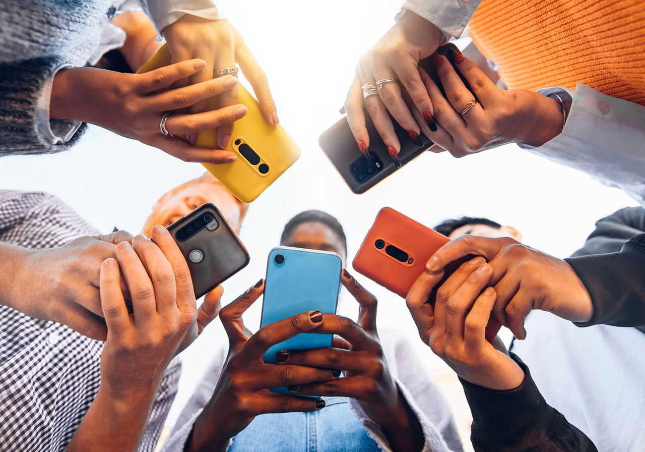 A circle of diverse people holding smartphones with colorful cases, viewed from below against a bright sky.