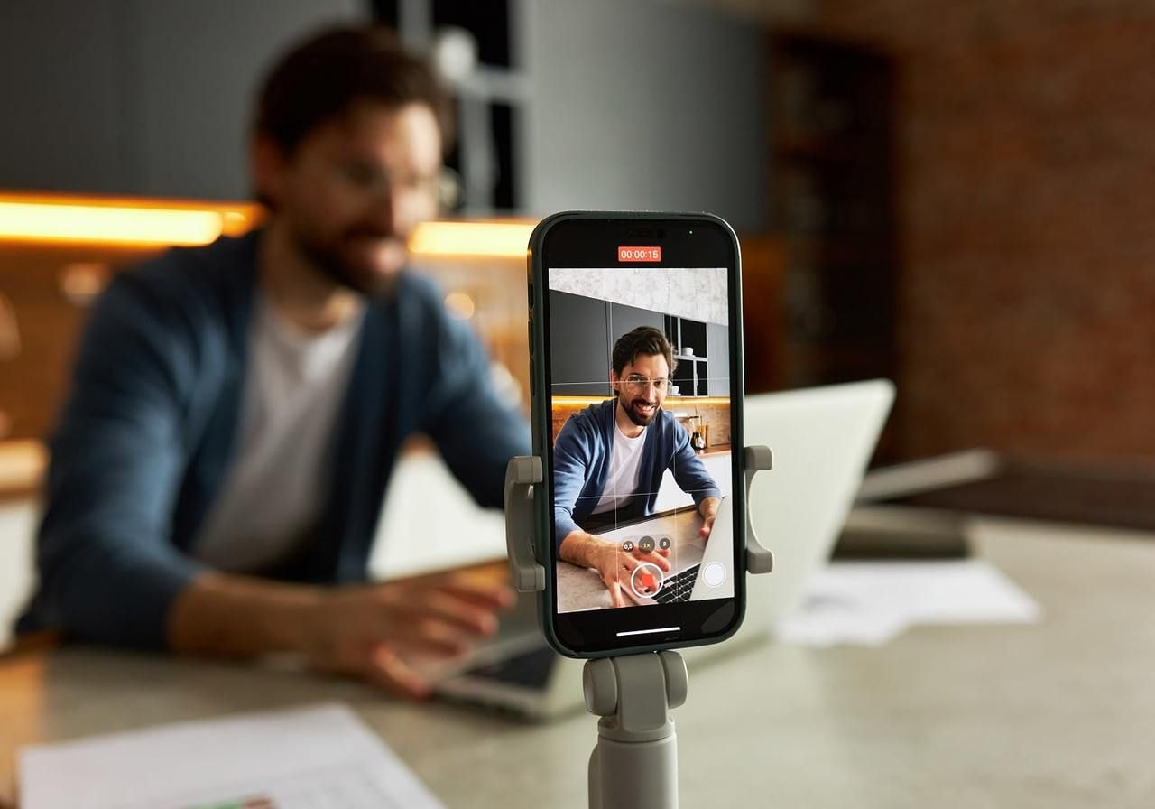 A bearded man in a blue sweater is smiling while sitting at a table with a laptop, recording himself with a phone.