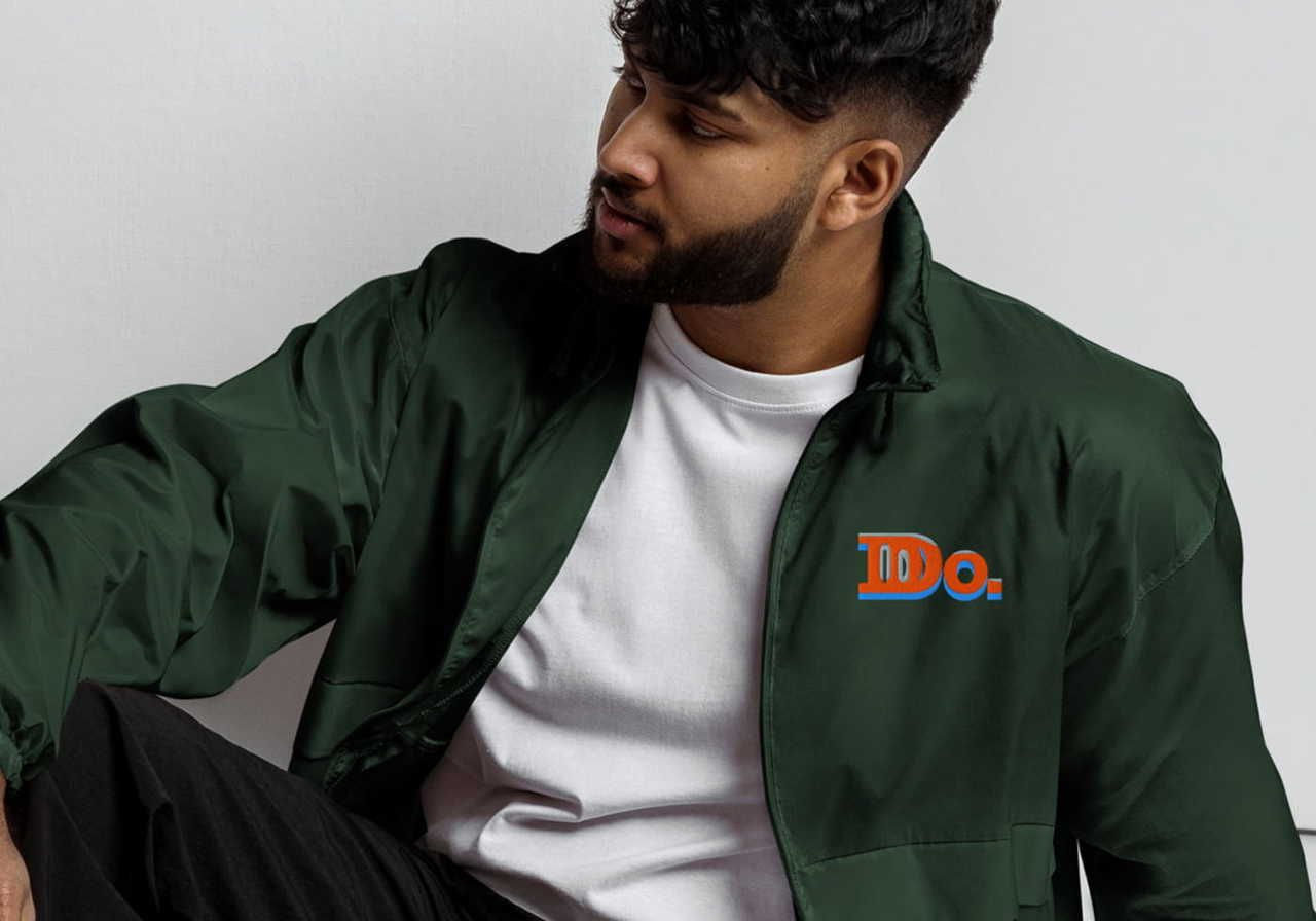 Man in a green jacket with "Do." logo, sitting casually, looking to the side. Wears a white t-shirt.
