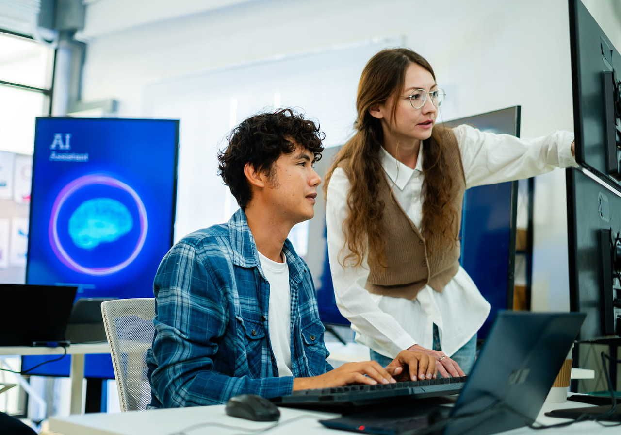 Two colleagues work at computers in a tech office with AI displays. The woman points at the screen while the man types.