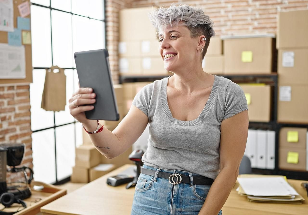 A woman in a gray shirt and jeans with short hair, smiling, holds a tablet in a bright office.