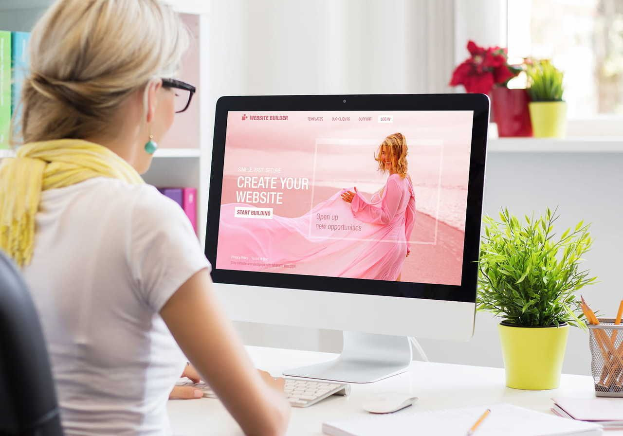 A woman at a desk looks at a computer screen displaying a website builder homepage featuring a woman in pink on a beach.