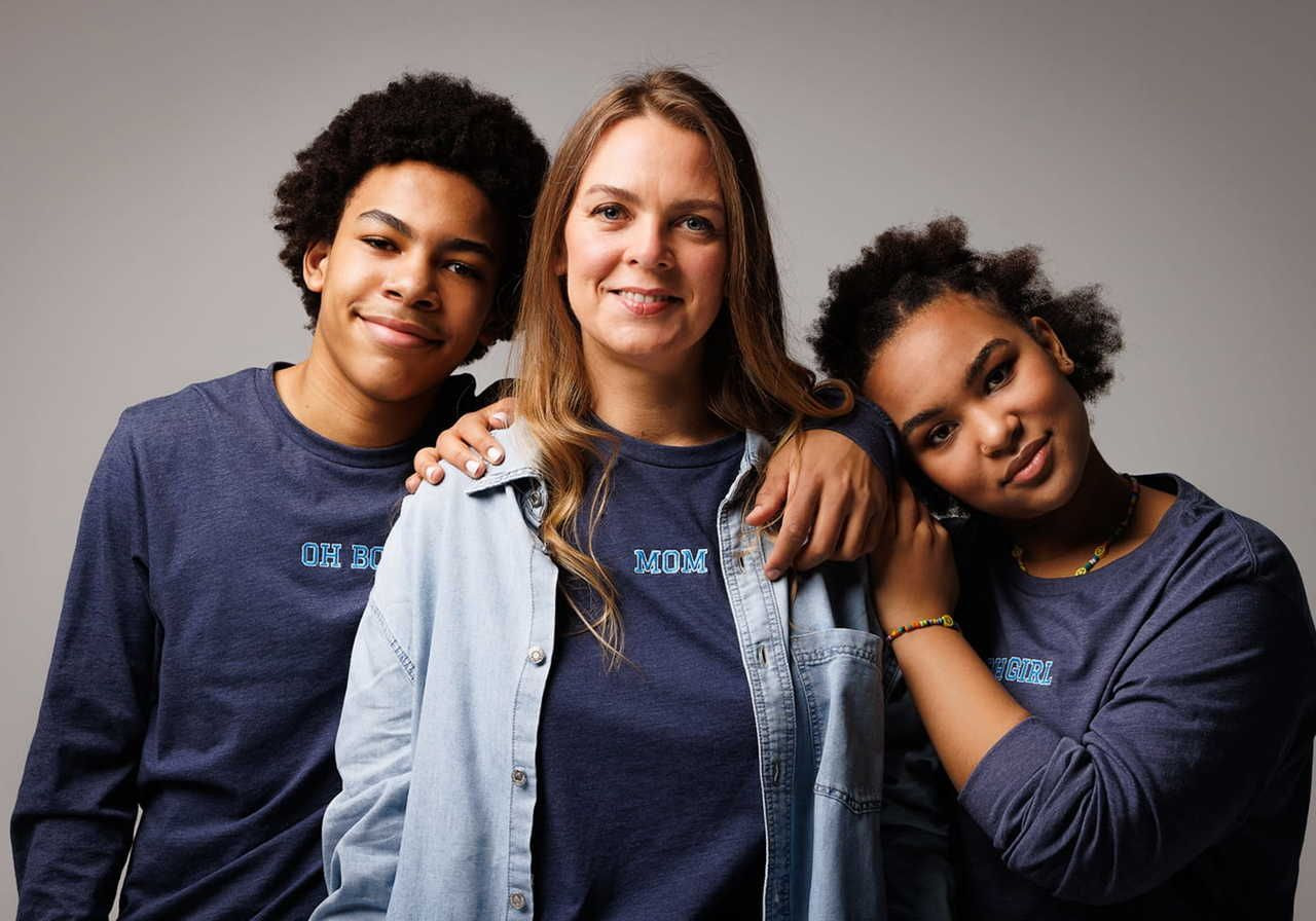 A woman and two teenagers smile together, with the woman standing in the middle in a denim jacket over her shirt. Teenagers wear matching blue shirts.
