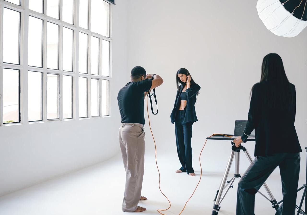 A photographer in a well-lit studio captures a model posing in a black outfit, while an assistant monitors a connected laptop.