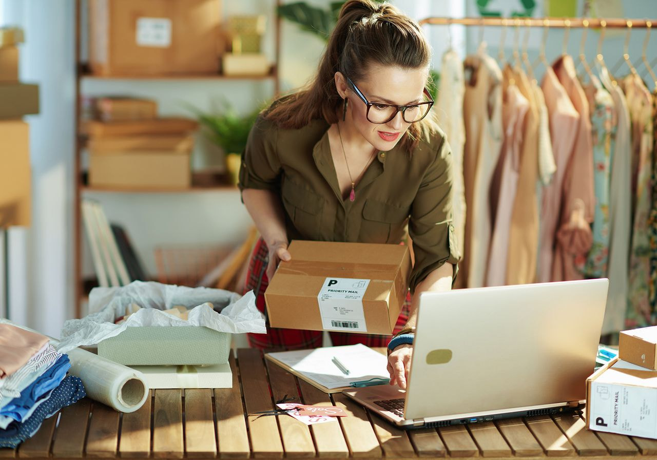 A woman works on her laptop while holding a box, focused on her task in a bright, organized workspace.
