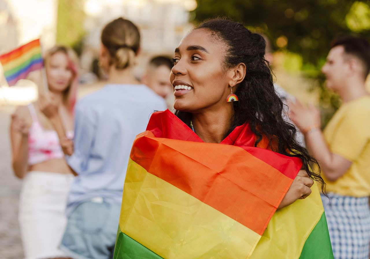 Smiling person wrapped in a rainbow flag, symbolizing LGBTQ+ pride. In the background, diverse group with rainbow flags, creating a joyful, inclusive atmosphere.