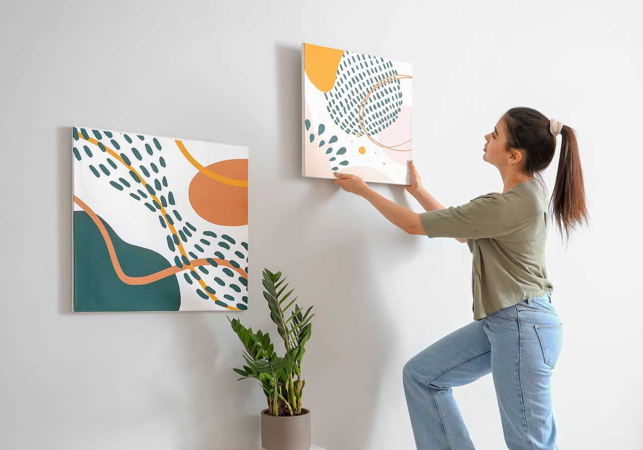 A woman hanging abstract artwork on a wall next to a potted plant.