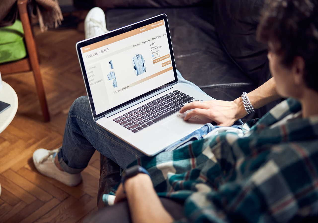 A man sits on a couch with a laptop, browsing an online shop displaying a blue shirt.