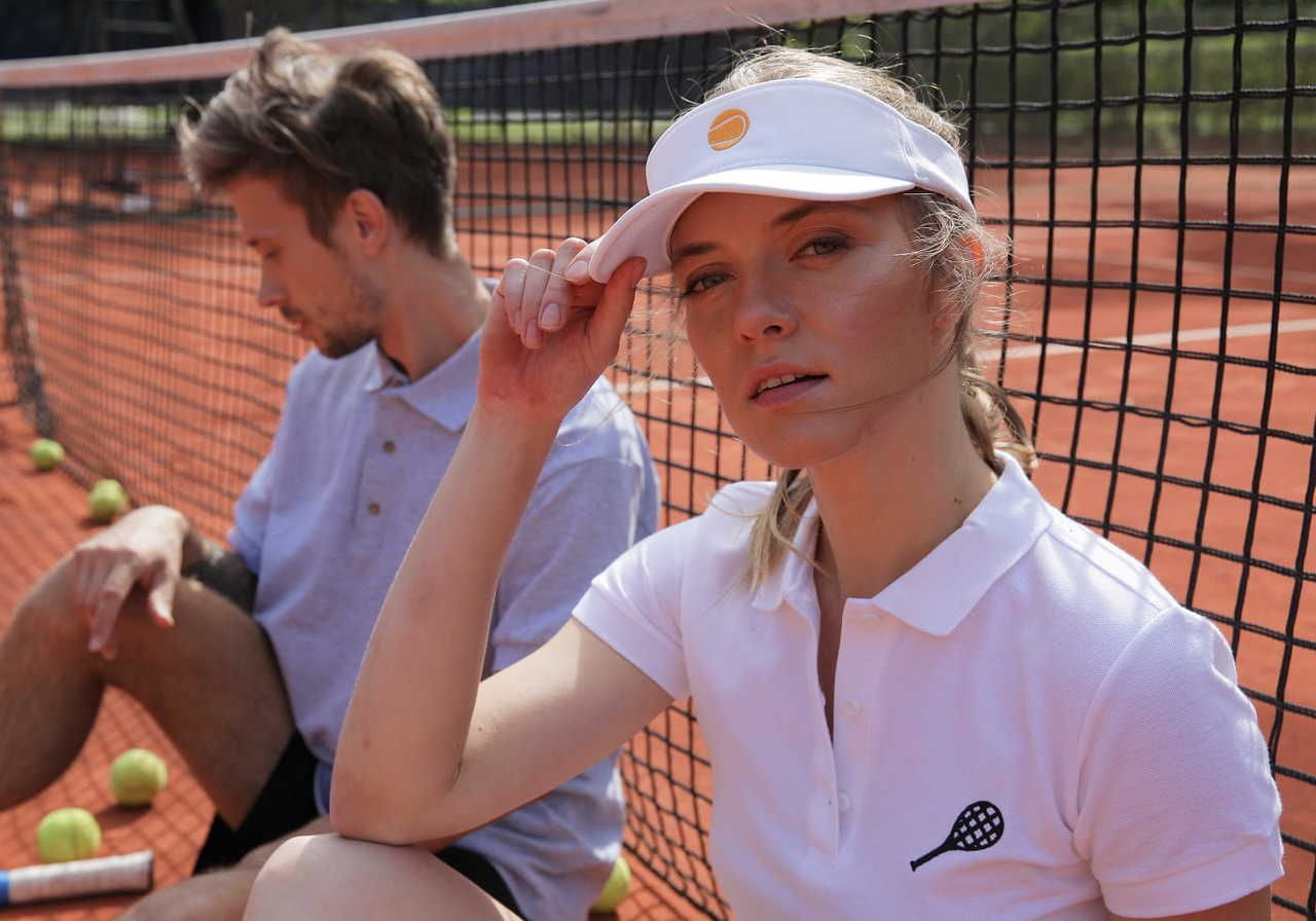 A man and a woman on a tennis court, with the woman wearing a white visor and a polo shirt, looking thoughtful while the man sits behind her.