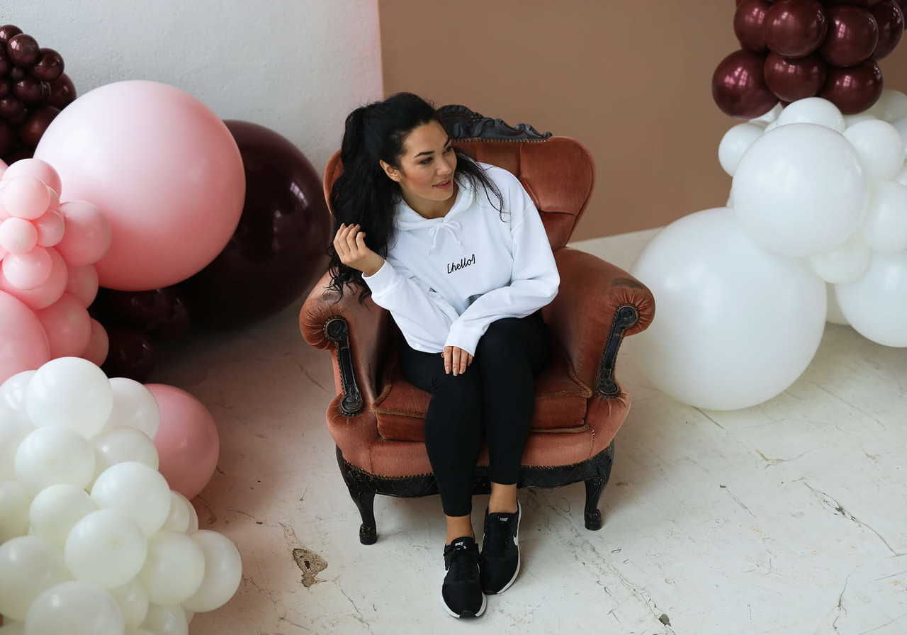 A woman sitting in a vintage armchair, wearing a white hoodie and black leggings, surrounded by pink and white balloons.