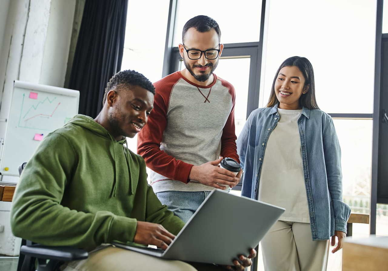 Three people engaged in a friendly office discussion around a laptop. One sits using the laptop, while the other two stand and smile.