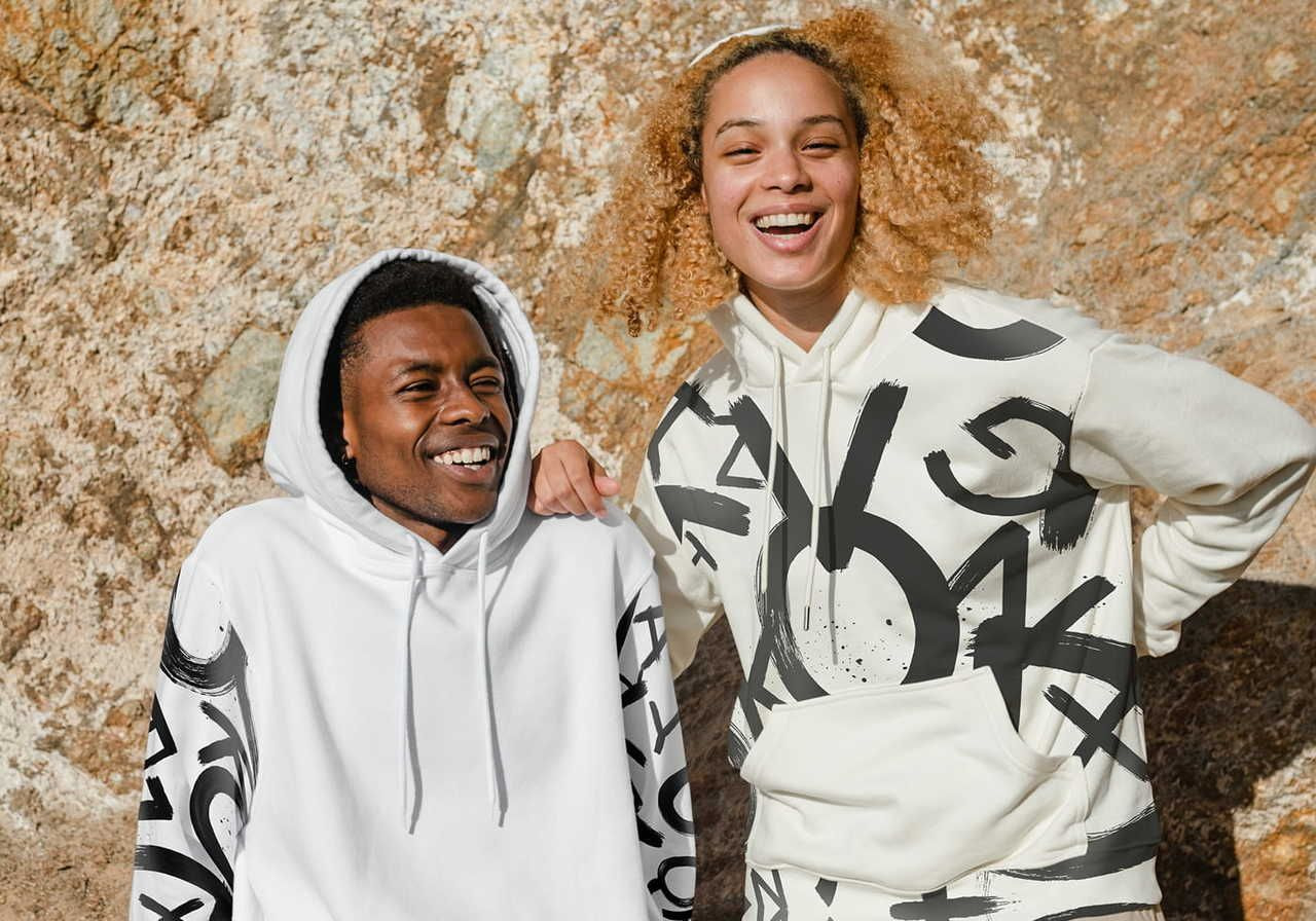 A smiling couple (man and woman), wearing white hoodies with abstract black prints against a stone background.