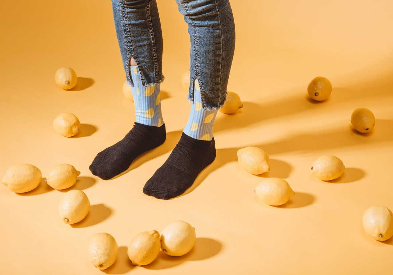 A person standing on a yellow background surrounded by lemons, wearing black socks and blue and yellow patterned socks, with distressed jeans.