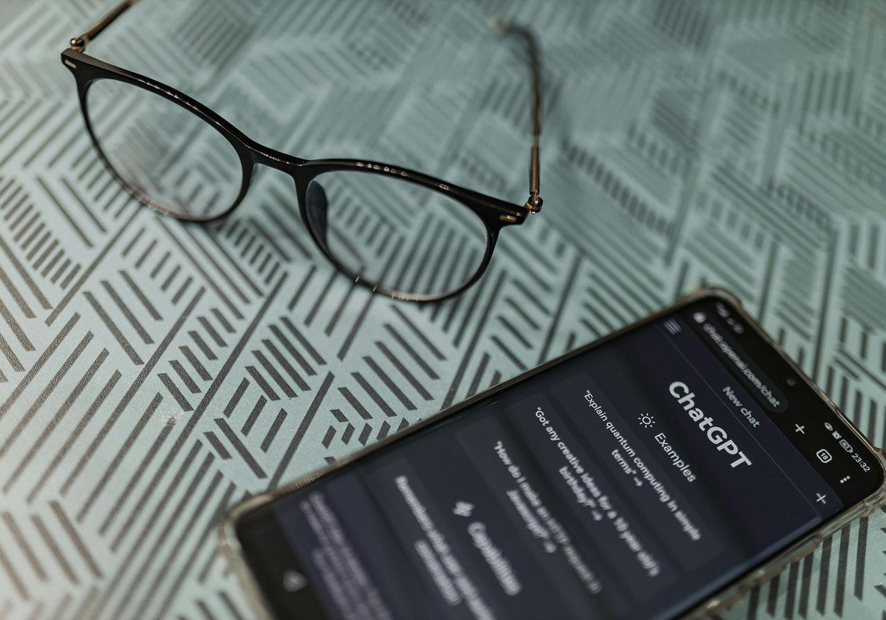 A smartphone and a pair of glasses resting on a wooden table surface.