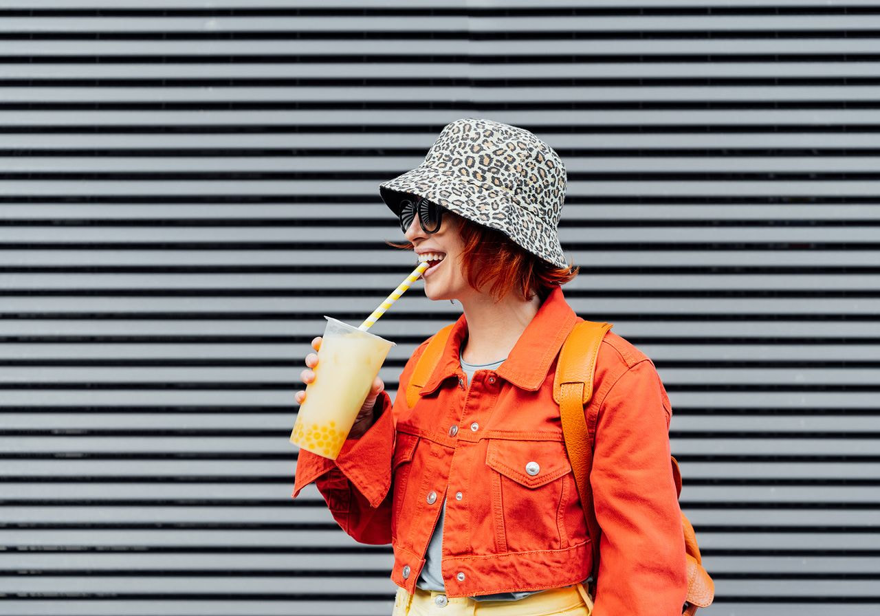A person in an orange jacket and leopard-print hat smiles while sipping bubble tea through a straw, standing against a striped gray background.