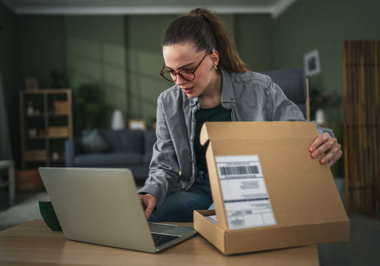 A woman is sitting at a table with a laptop open in front of her, looking at a package with a shipping label in her hands.