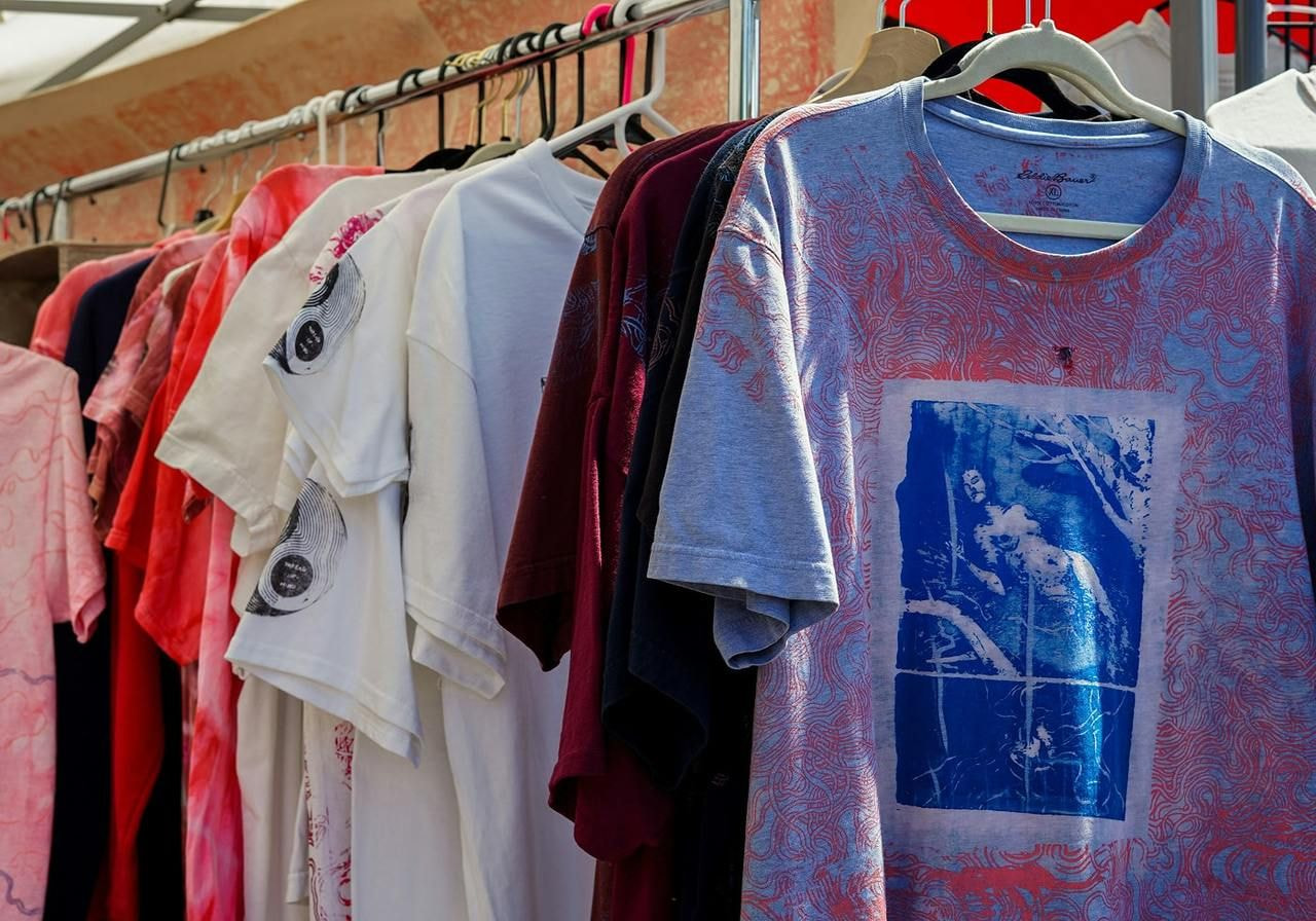 A rack of colorful t-shirts on hangers, featuring various designs. The foreground shirt is light blue with an abstract retro-wave print.