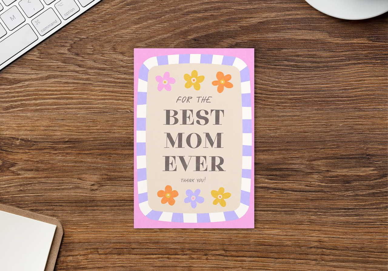 A card on a wooden desk reads "For the Best Mom Ever" with colorful flowers and a striped border.