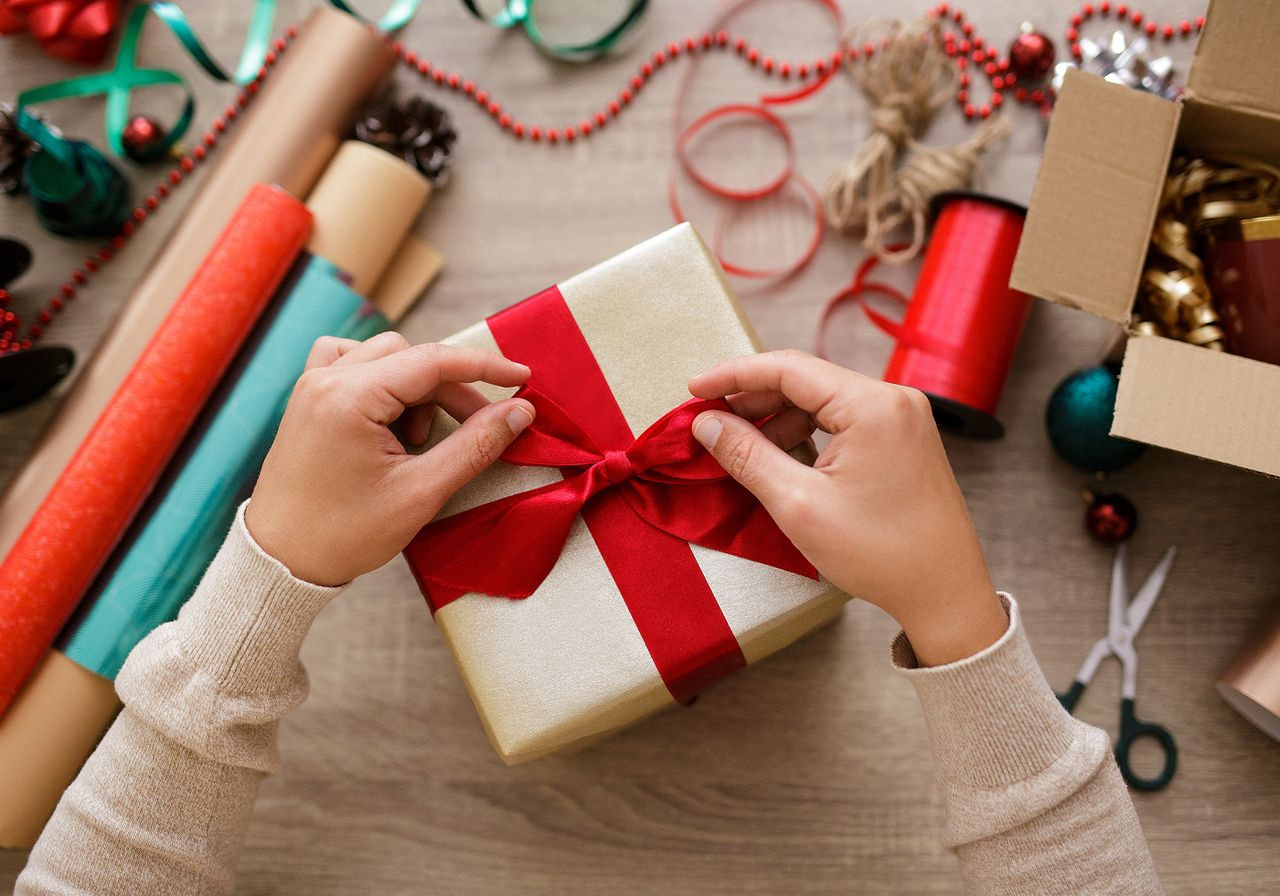 Hands tie a red satin bow on a gold-wrapped gift, surrounded by rolls of wrapping paper, ribbon spools, twine, and Christmas ornaments.