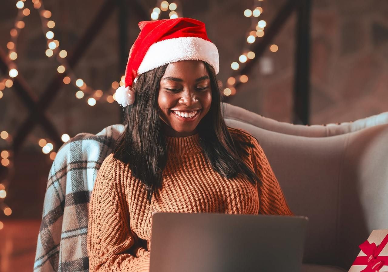 A woman in a Santa hat smiles while using a laptop, sitting on a cozy sofa. Warm fairy lights in the background.