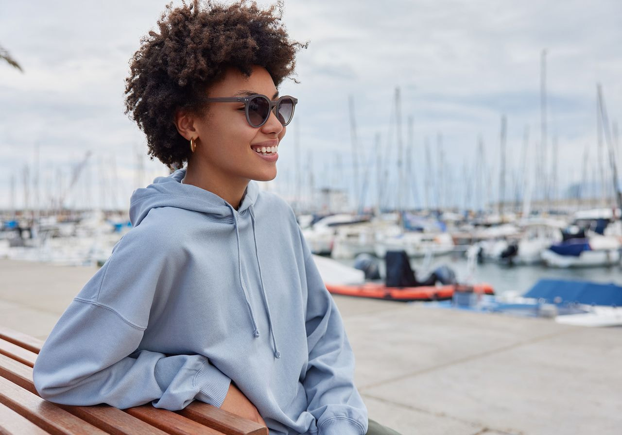 A smiling woman in sunglasses and a light blue oversized hoodie sits on a wooden bench at a marina with sailboats in the background.