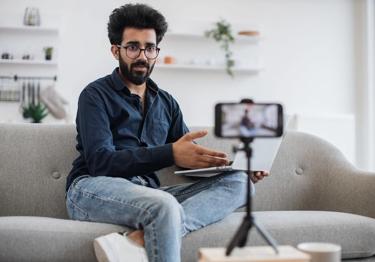A man with glasses sits on a gray sofa, holding a laptop, gesturing while recording a video on a smartphone.