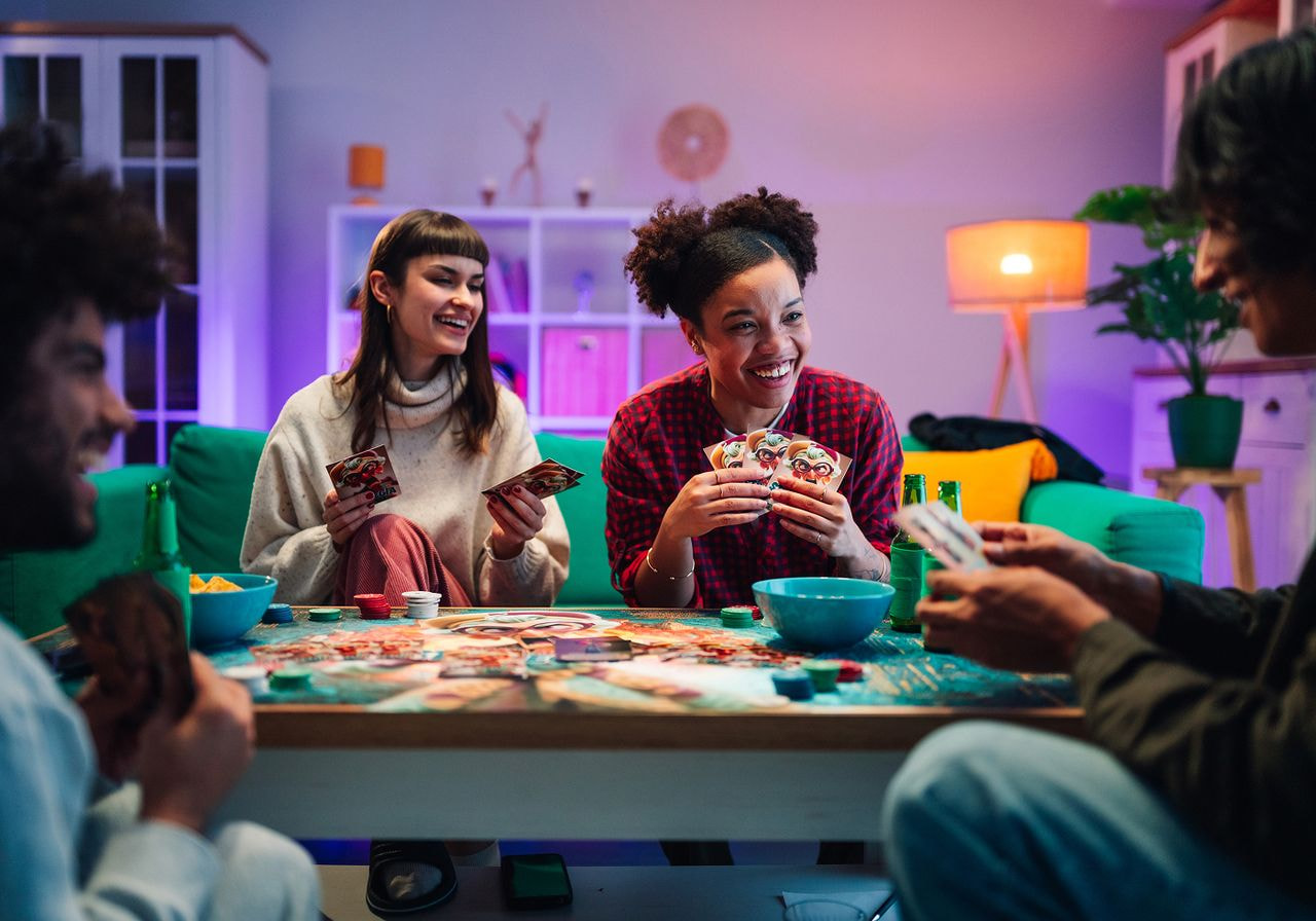 Four friends gathered in a living room, engaged in a lively board game, with laughter and excitement visible on their faces.