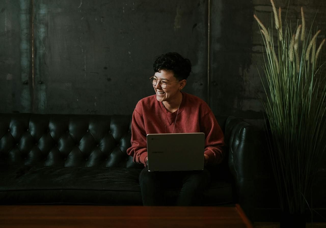 A woman sitting on a couch, focused on her laptop, with a cozy living room setting in the background.