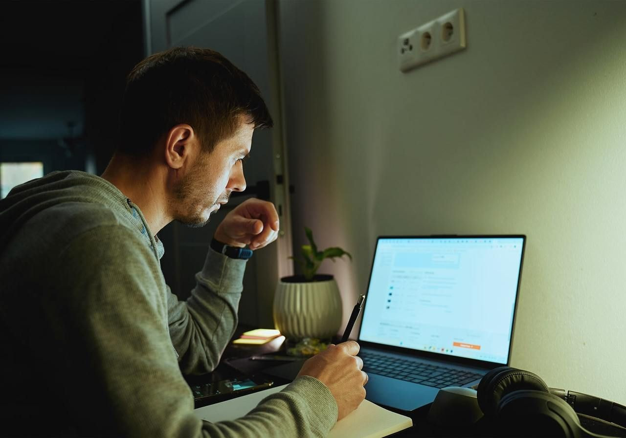 A man in a hoodie is using a laptop to find out how to start a hoodie business. A potted plant and headphones are nearby.