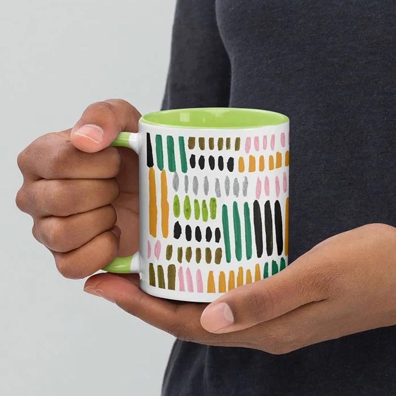 Person holding a mug with a colorful pattern of vertical dashes in green, orange, pink, black, and gray.