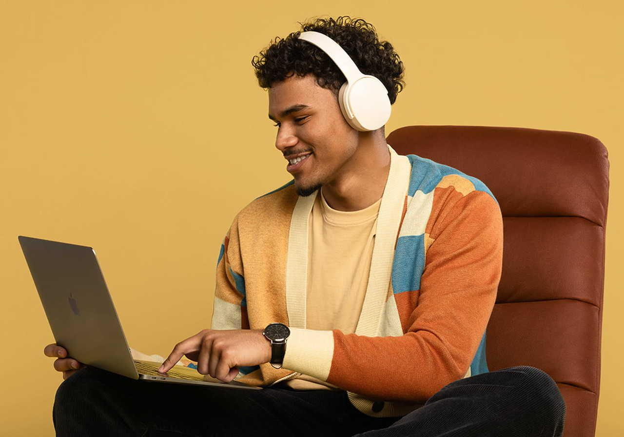 A young man in a colorful cardigan and headphones smiles while using a laptop. He's relaxed in a brown chair, against a yellow background.
