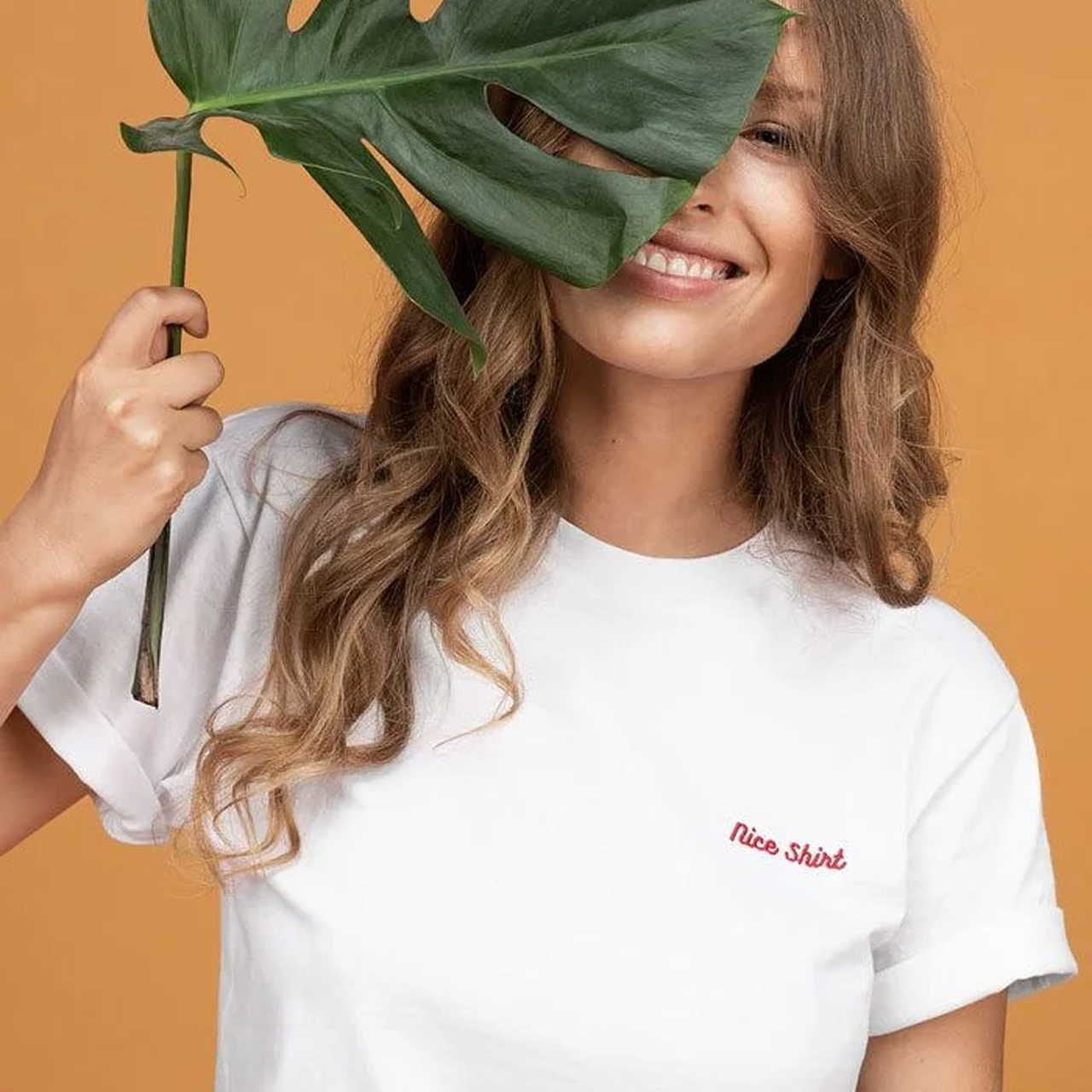 A woman is holding a green leaf, wearing a white Unisex Staple T-Shirt | Bella + Canvas 3001 with “nice” embroidered in red.
