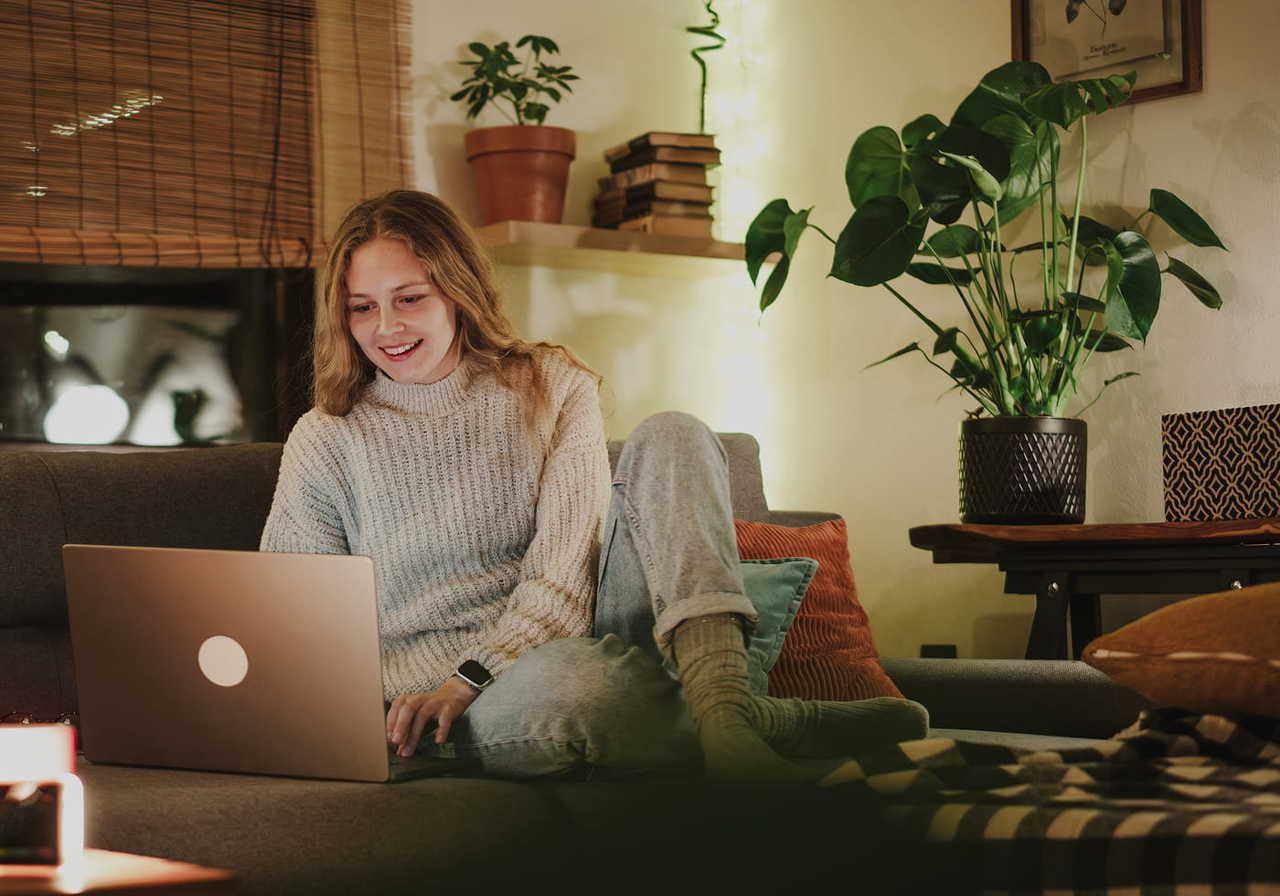Young woman in cozy sweater, smiling at laptop on couch, surrounded by plants and soft lighting.