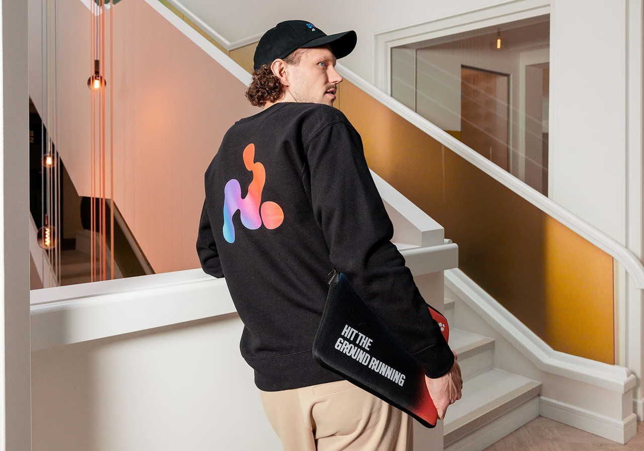 A man wearing a black sweatshirt with a colorful logo on the back, standing near a staircase and holding a bag that says 