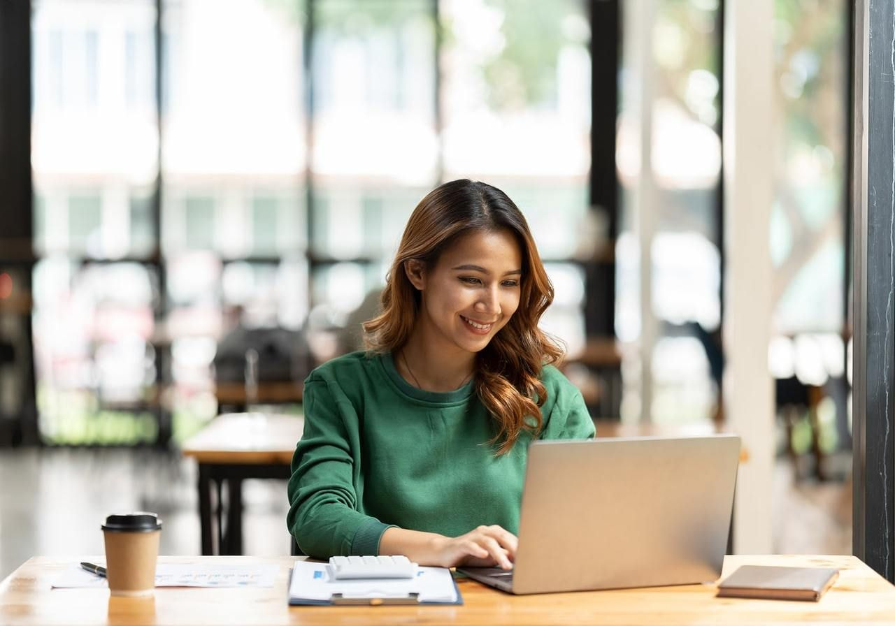 A smiling woman is sitting at a table and using a laptop to find out how to start a greeting card business.