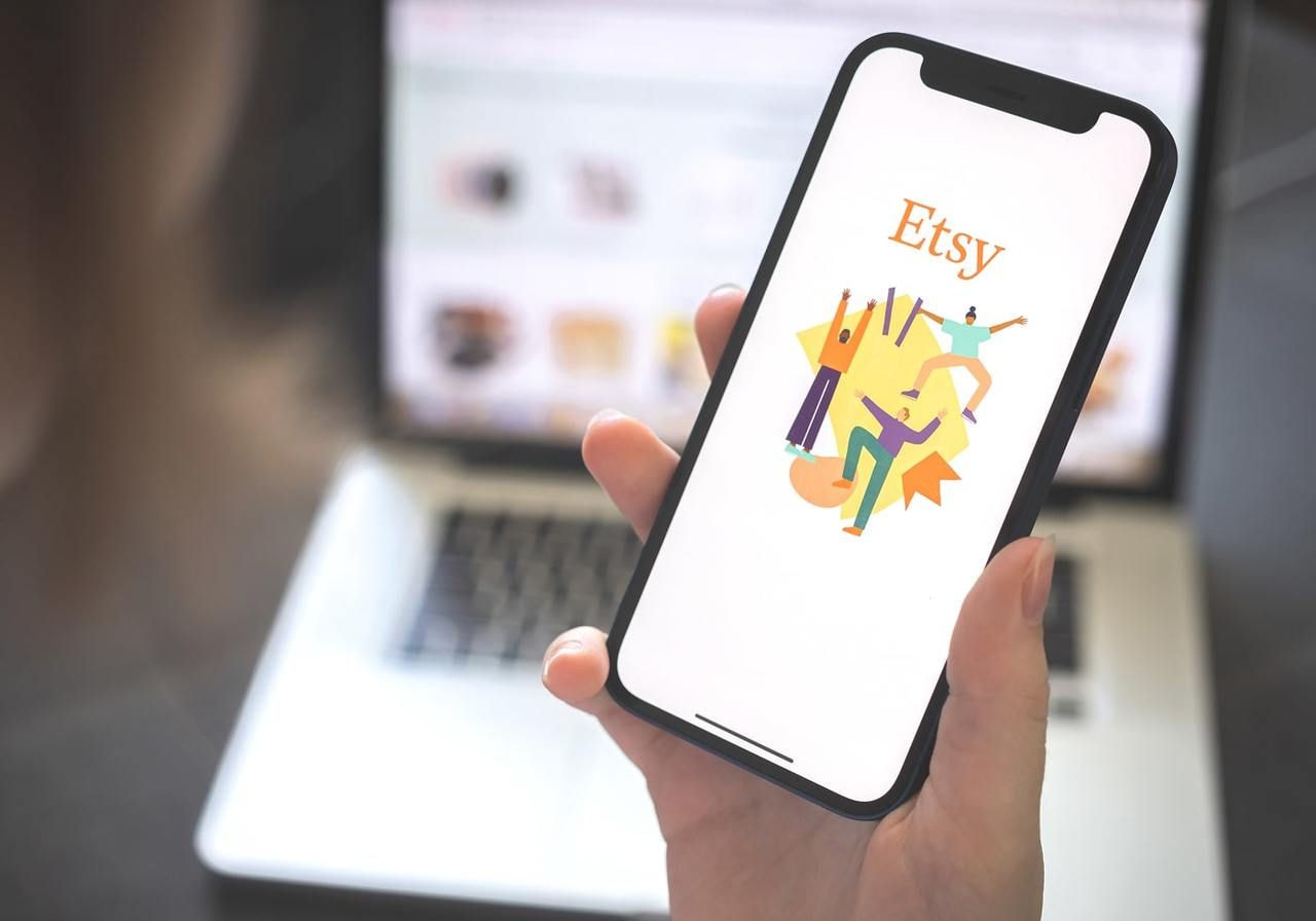 A hand holds a smartphone displaying the Etsy app with colorful abstract art. A blurred laptop in the background.