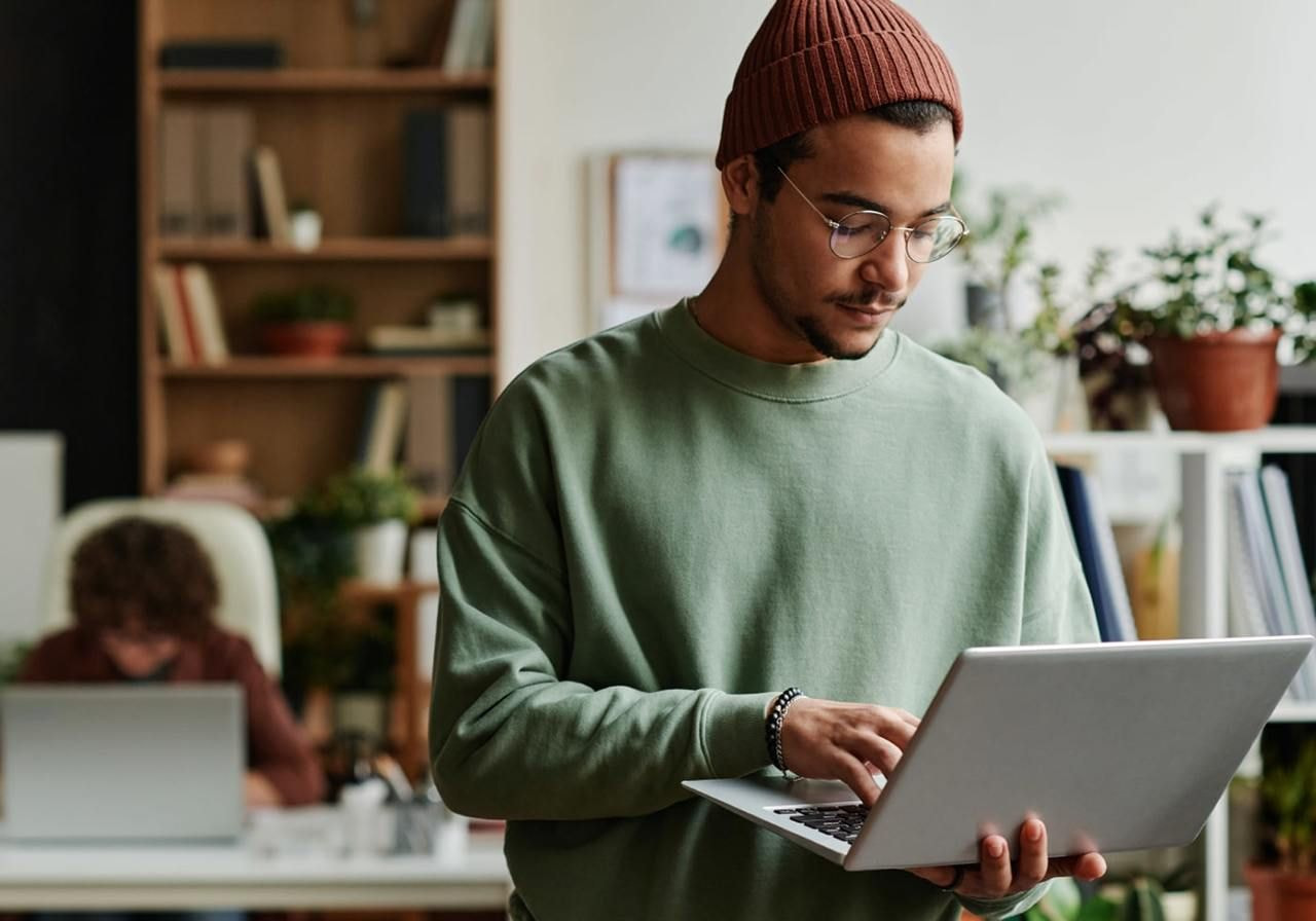 A man in a green sweater and a brown beanie works on a laptop in a cozy office. The background shows bookshelves and plants.