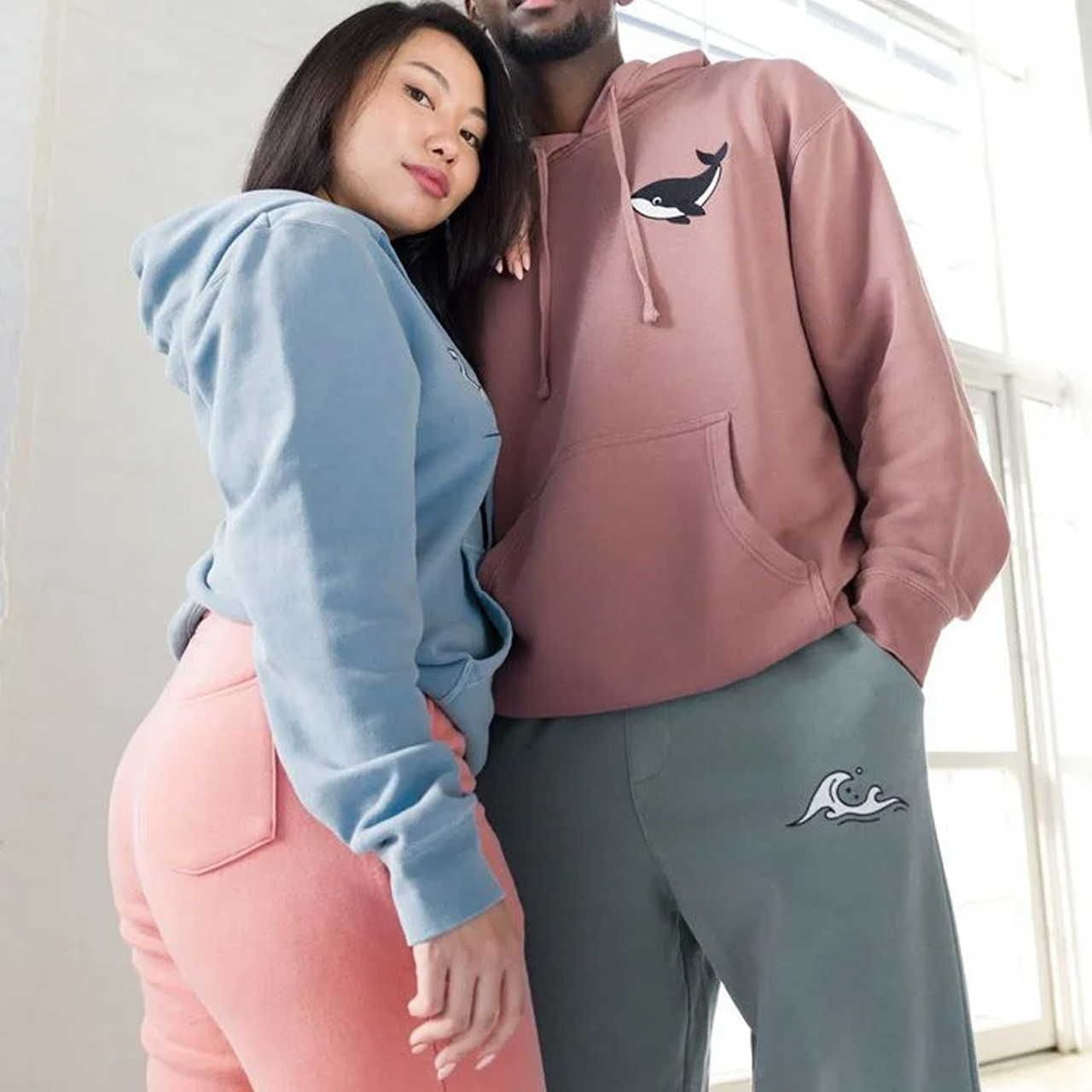 Two people wearing hoodies stand together. The woman wears a blue hoodie and pink pants; the man wears a brown hoodie with a whale design.