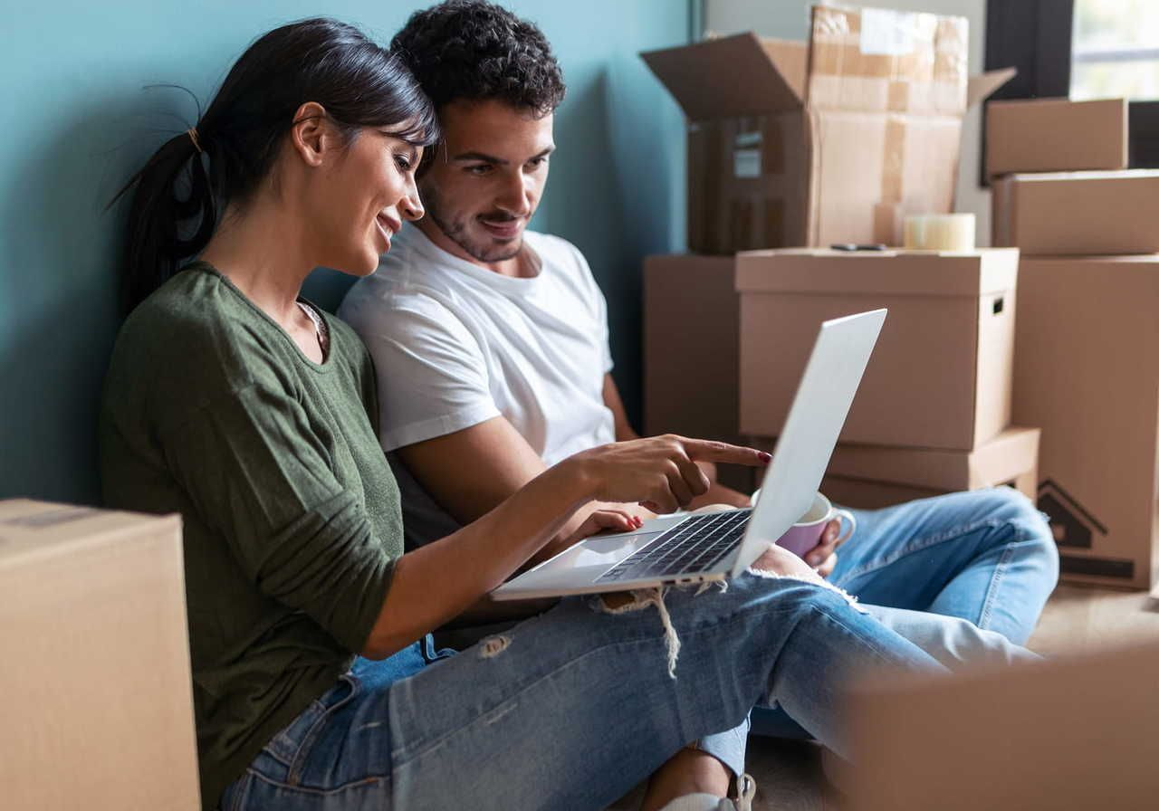 A couple sitting on the floor surrounded by moving boxes, using a laptop and smiling while pointing at the screen.