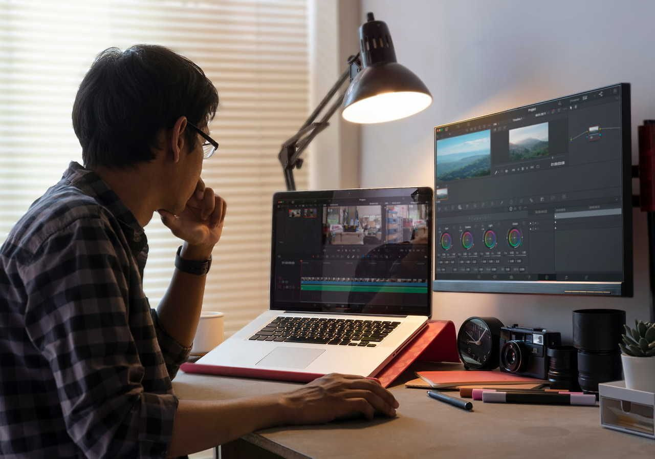 A man working on video editing software on a laptop at a desk, with a large monitor displaying editing timelines, various camera equipment nearby.
