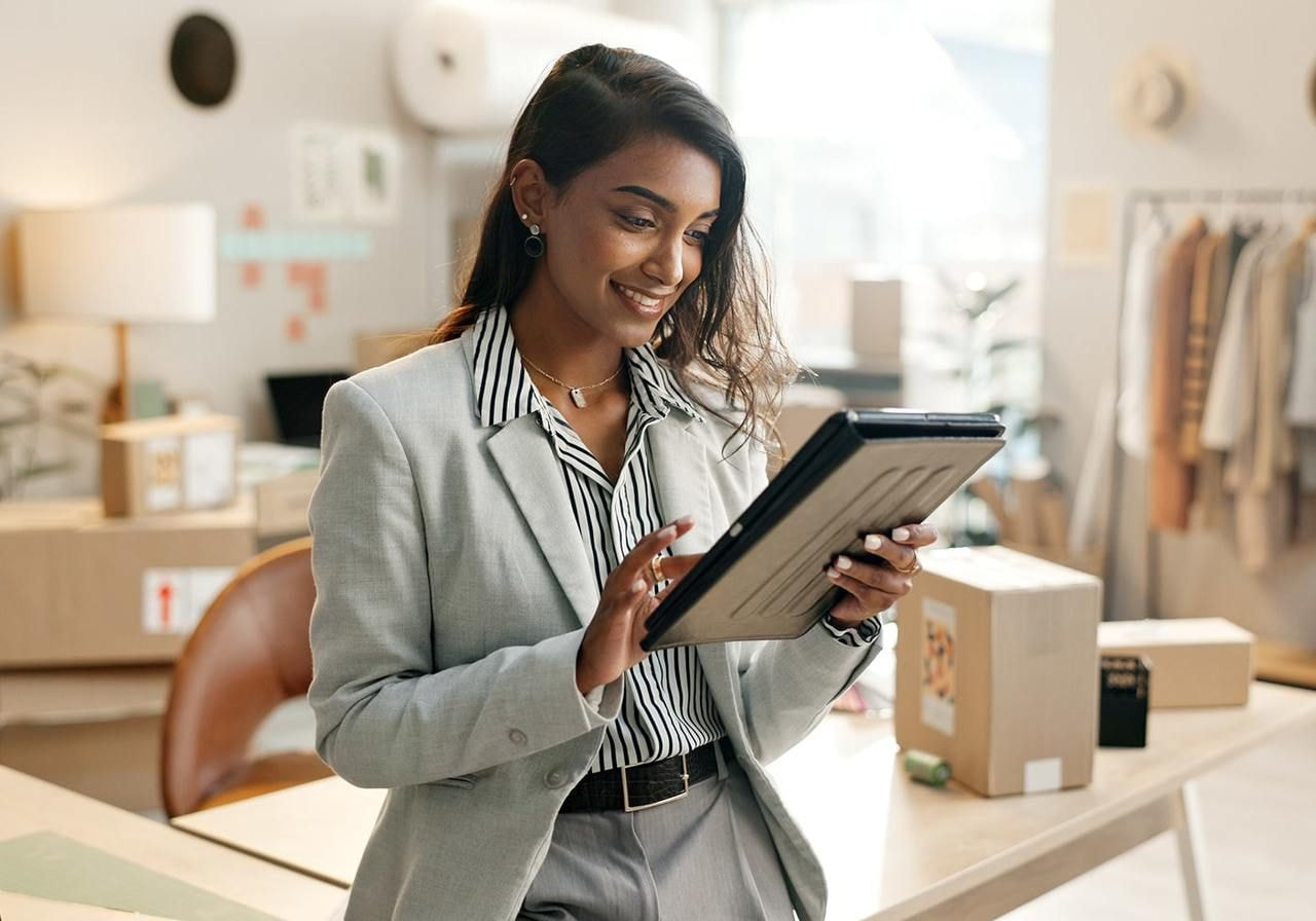 A smiling woman in a business suit stands in an office while using a tablet to find out what print-on-demand is.