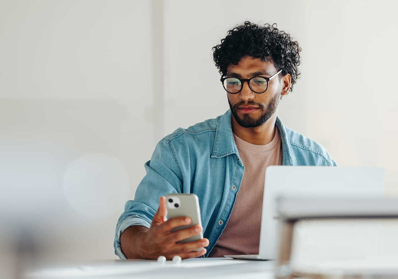 A man with curly hair and glasses sits at a desk, looking at his smartphone. He wears a denim shirt over a pink t-shirt, with a laptop open nearby.
