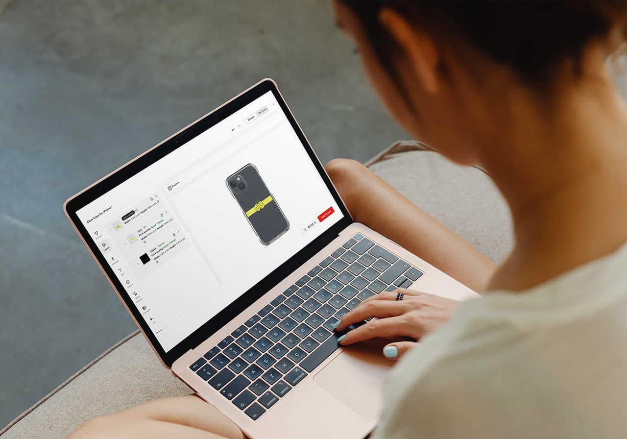 A person is using a laptop, designing a custom phone case cover using Printful’s Design Maker.