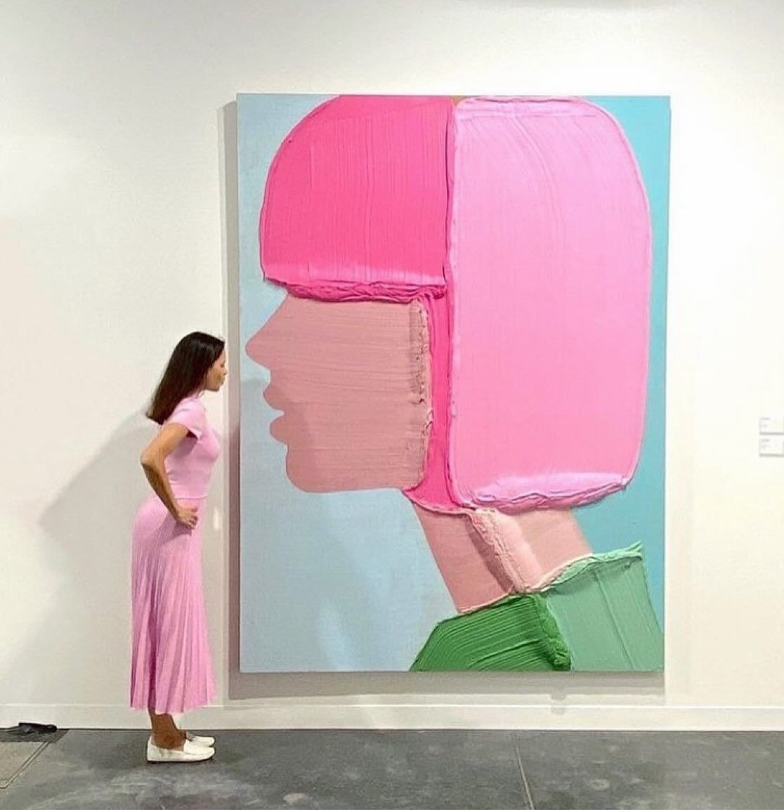 A woman in a pink dress stands beside a large, colorful painting in an art gallery setting.