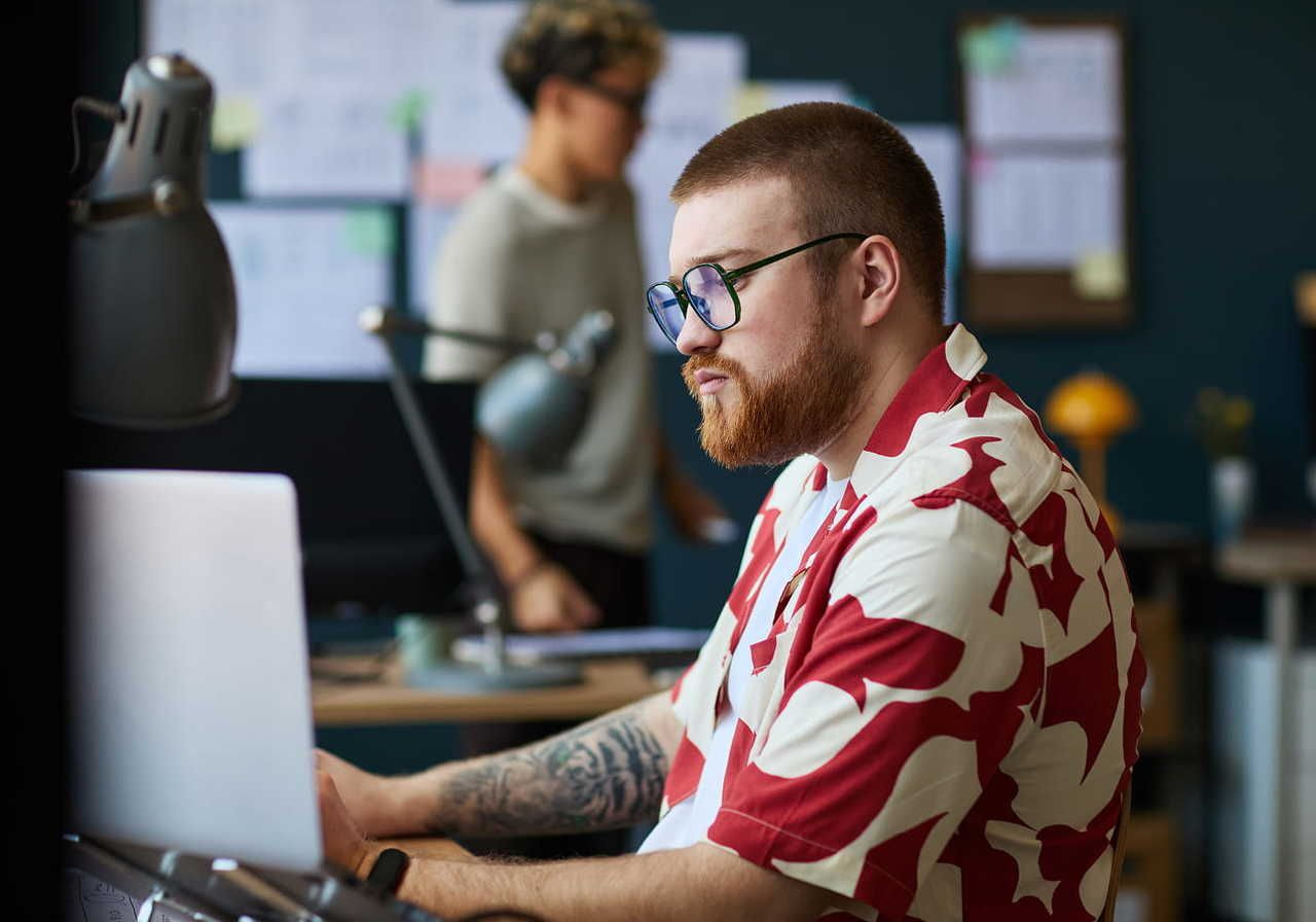 A man with glasses and a patterned shirt focuses on his laptop, understanding the difference between RGB and CMYK.