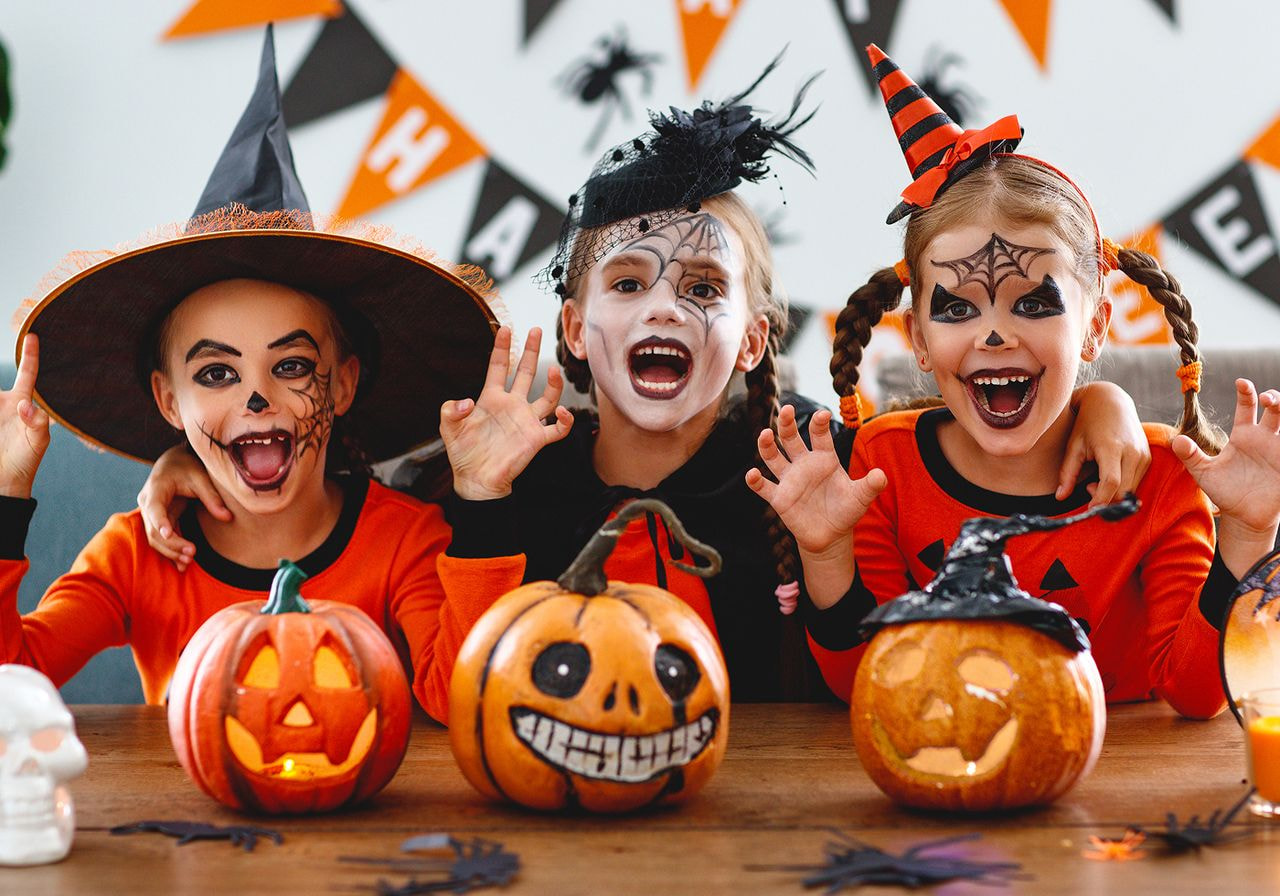 Three children in Halloween costumes and face paint pose playfully with carved pumpkins. 