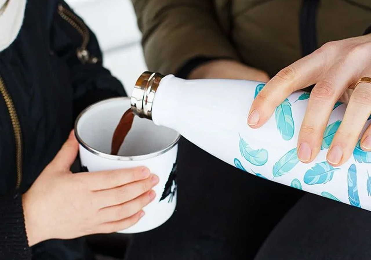 A person pours a warm drink from a white bottle with blue feathers into a mug held by a child.